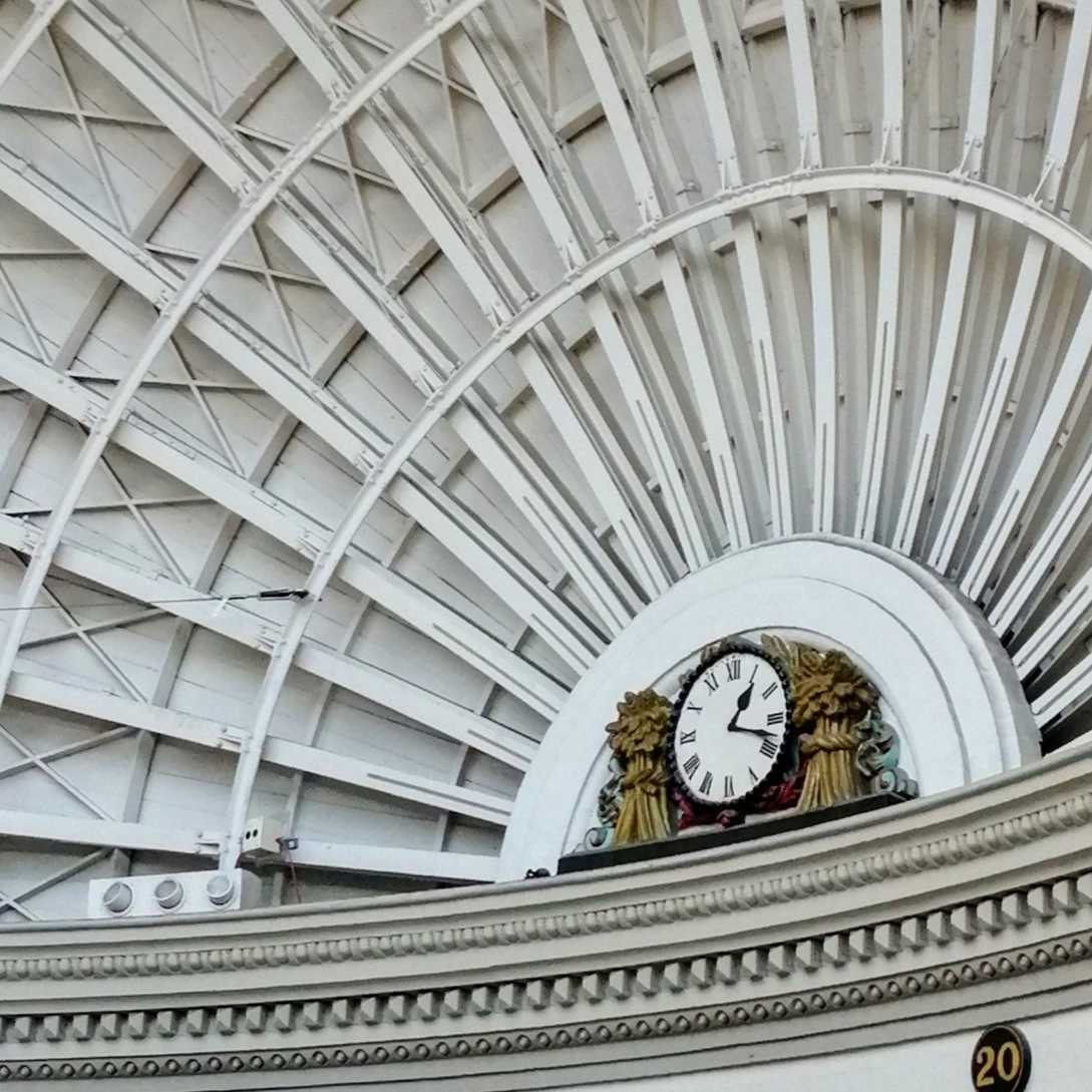 Photograph of an analog clock, on either side there are gold bundles of corn stood upright, the clock is set into the white roof of a domed building.