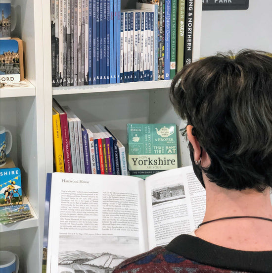 Photograph of bookshelves holding books on Yorkshire, a person with short hair is stood in front of the shelf to the right holding open a book on a page about Harewood House.